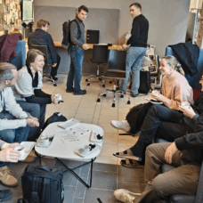 Suunnitteluporukoita keskustelemassa kahdella vastakkaisella korkeaselkänojaisella sohvalla, välissä pyöreä pöytä. Taustalla kaksi suunnittelijaa katsomassa pöydässä keskustelijoita.