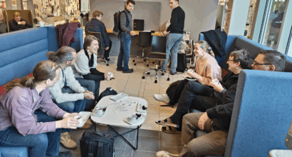 Suunnitteluporukoita keskustelemassa kahdella vastakkaisella korkeaselkänojaisella sohvalla, välissä pyöreä pöytä. Taustalla kaksi suunnittelijaa katsomassa pöydässä keskustelijoita.