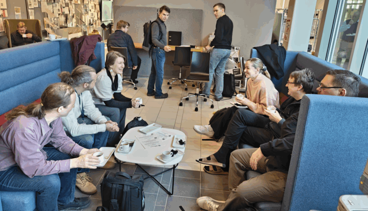 Suunnitteluporukoita keskustelemassa kahdella vastakkaisella korkeaselkänojaisella sohvalla, välissä pyöreä pöytä. Taustalla kaksi suunnittelijaa katsomassa pöydässä keskustelijoita.