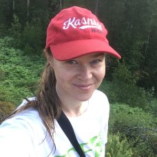 Jonna Kukkonen katsoo kameraan ja hymyilee lähikuvassa lippis päässä metsässä.