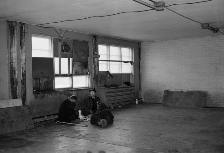 Mustavalkoinen kuva, jossa kolme ihmistä istuu hylätyn talon lattialla.