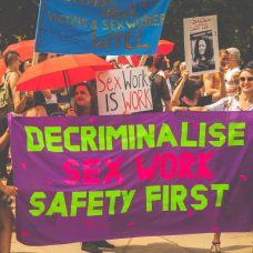 Mielenosoittajia kadulla, edessä kangas, jossa lukee "decriminalise sex work - safety first".