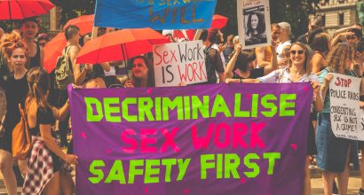 Mielenosoittajia kadulla, edessä kangas, jossa lukee "decriminalise sex work - safety first".