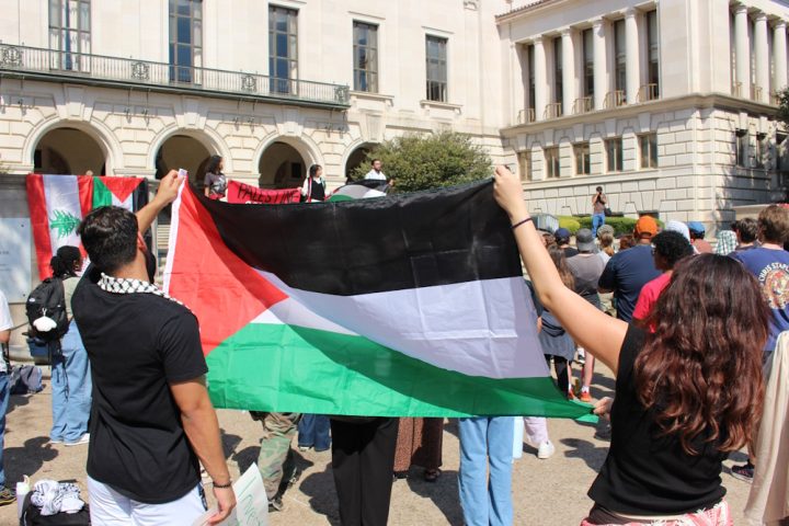 Ihmisiä kokoontuneena ulos rakennuksen eteen, kuvan edustalla kaksi henkilä pitelee Palestiinan lippua ja taustalla näkyy myös Palestiinan lippuja.