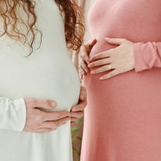 Kaksi raskaana olevaa henkilöä seisoo vierekkäin ja pitelee vatsojaan, toisella valkoinen ja toisella vaaleanpunainen mekko. Vain keskivartalot näkyvät.