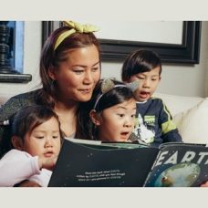 Aikuinen ja kolme lasta istuvat sohvalla ja katsovat kirjaa.