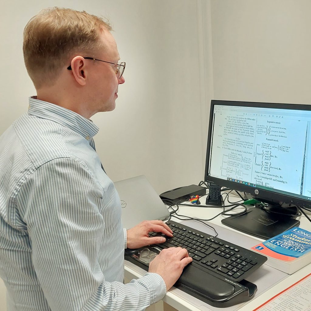 Side profile view of Dr Janne Skaffari looking at a 17th-century grammar on a computer screen, with a printed 21st-century Longman grammar book on his desk.