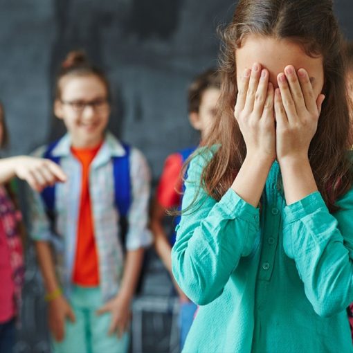 Tyttö seisoo koululuokassa kädet silmillä, takana on neljä lasta, joista yksi osoittaa häntä sormella