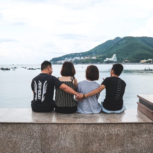 Neljä ihmistä istuu selkä kuvaajaan päin meren äärellä, laitimmaiset pitävät toisiaan selän takaa kädestä kiinni.