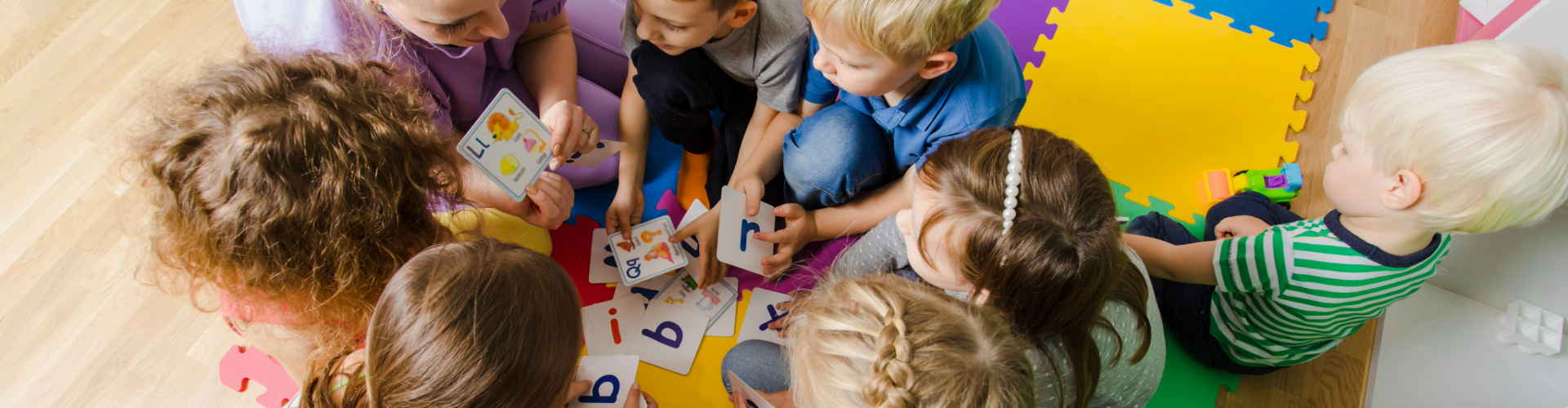 Aikuinen ja usemapi lapsi kuvattuna yläviistosta pelaamassa korttia värikkäälä alustalla.