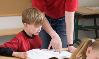 Lapsi istuu pulpetissa, opettaja näyttää hänelle oppikirjasta jotain.