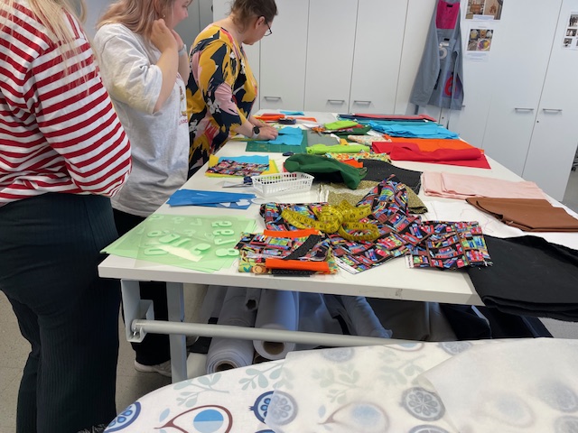 Kolme ihmistä tekee käsitöitä värikkäiden kankaiden parissa.