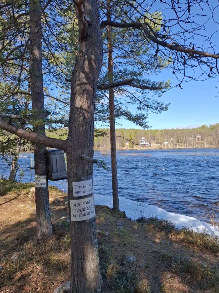 Kuva puusta joen varrella. Puuhun kiinnitetty kyltit, joissa lukee "This in not Juutua trail" ja "Not for tourist! Ei turisteille".