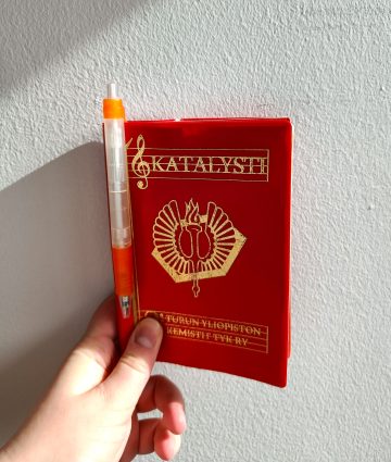 Kuva Turun yliopiston kemistien TYK ryn laulukirjasta, Katalystistä. Kirja on kirkkaan punainen ja siinä on Loimun oranssi kynä kiinni. Kannessa on TYK ryn logo kultaisella. 
