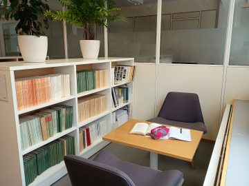 Kuvassa kaksi nojatuolia työpöydän ääressä. Toisella puolell apöytää on ikkuna ja toisella matala kirjahylly, jonka päällä on kaksi elävää kasvia.