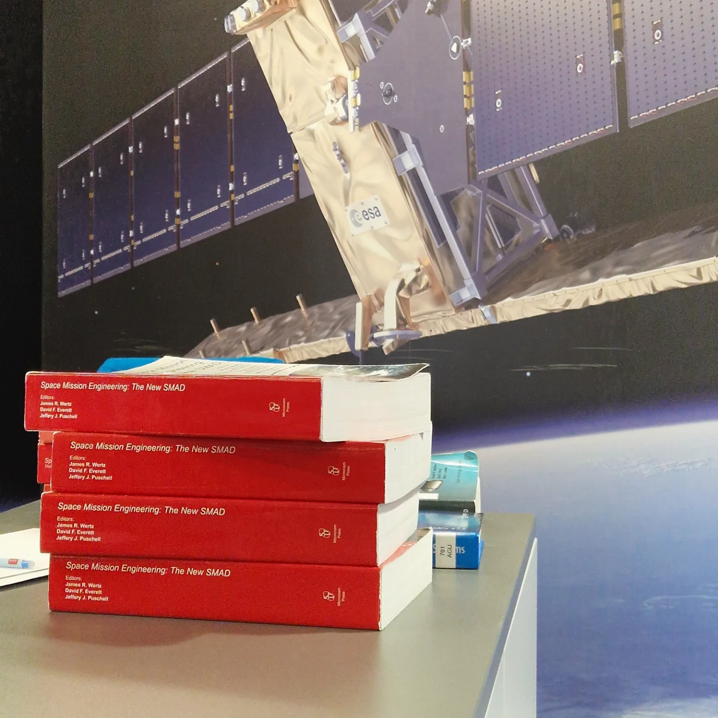 Kasa punaisia oppikirjoja pinossa pöydällä. Kirjojen selkämyksessä lukee "Space Mission Engineering: the new SMAD". Taustalla seinä, jossa on iso kuva satelliitista. 