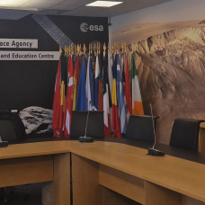 Euroopan avaruusjärjestön avaruusturvallisuus- ja koulutuskeskuksen kokoushuoneesta, kuvassa puinen tyhjä koferenssipöytä, taustalla eri maiden lippuja ja seinällä kuvia avaruudesta ja teksti "European Space Agency, Space Security and Education Centre".