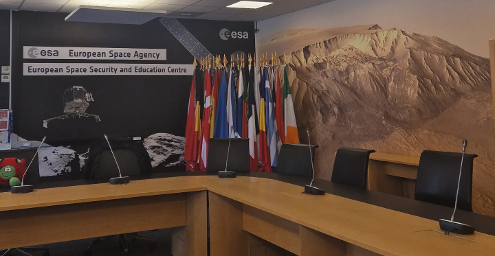 Euroopan avaruusjärjestön avaruusturvallisuus- ja koulutuskeskuksen kokoushuoneesta, kuvassa puinen tyhjä koferenssipöytä, taustalla eri maiden lippuja ja seinällä kuvia avaruudesta ja teksti "European Space Agency, Space Security and Education Centre".