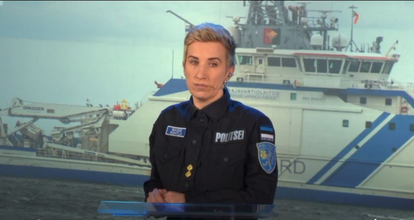 A woman in a blue uniform standing in front of a picture of white and blue a Finnish Border Guard vessel.