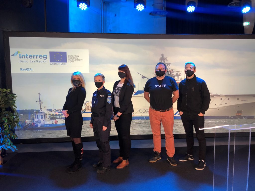 Five persons with black facial masks posing in front of a picture of two patrol vessels in a room lit by blue light.