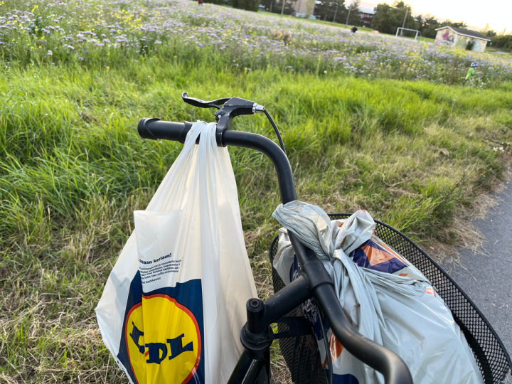A picture of a bicycle with a plastic bag full with groceries in a bicycle basket and a plastic bag full with groceries on a handlebar.