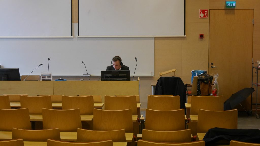 Väittelijä istuu tietokoneen ääressä kuulokkeet korvilla tyhjän luentosalin etuosassa.