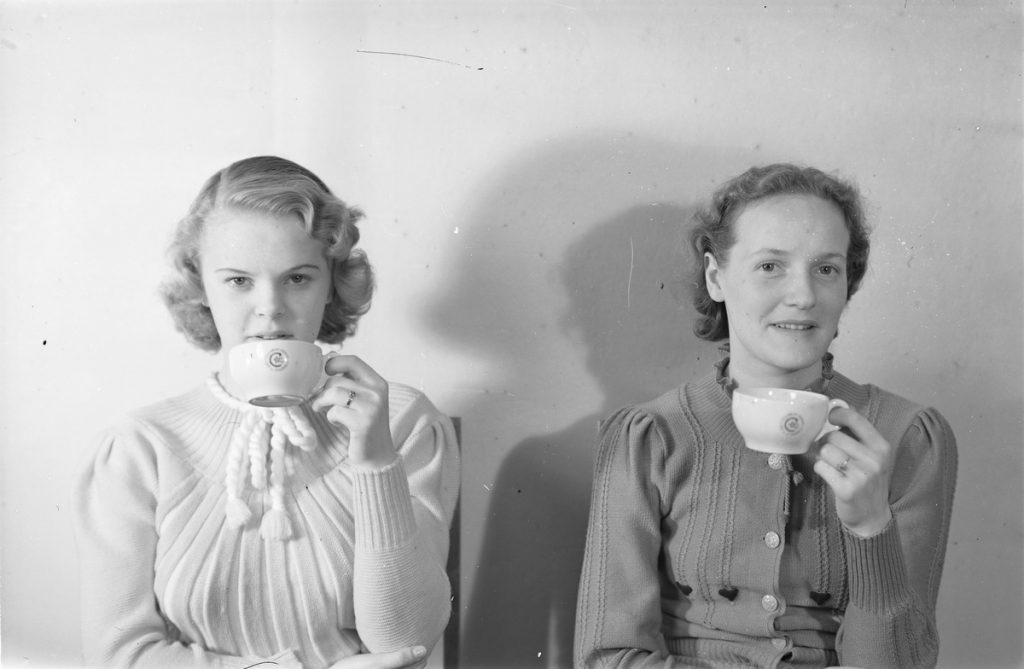 Kaksi naista juovat kahvia. Molemmilla on lyhyet, kiharat hiukset ja valkoinen kahvikuppi vasemmassa kädessään. He katsovat suoraan kameraan ja hymyilevät.