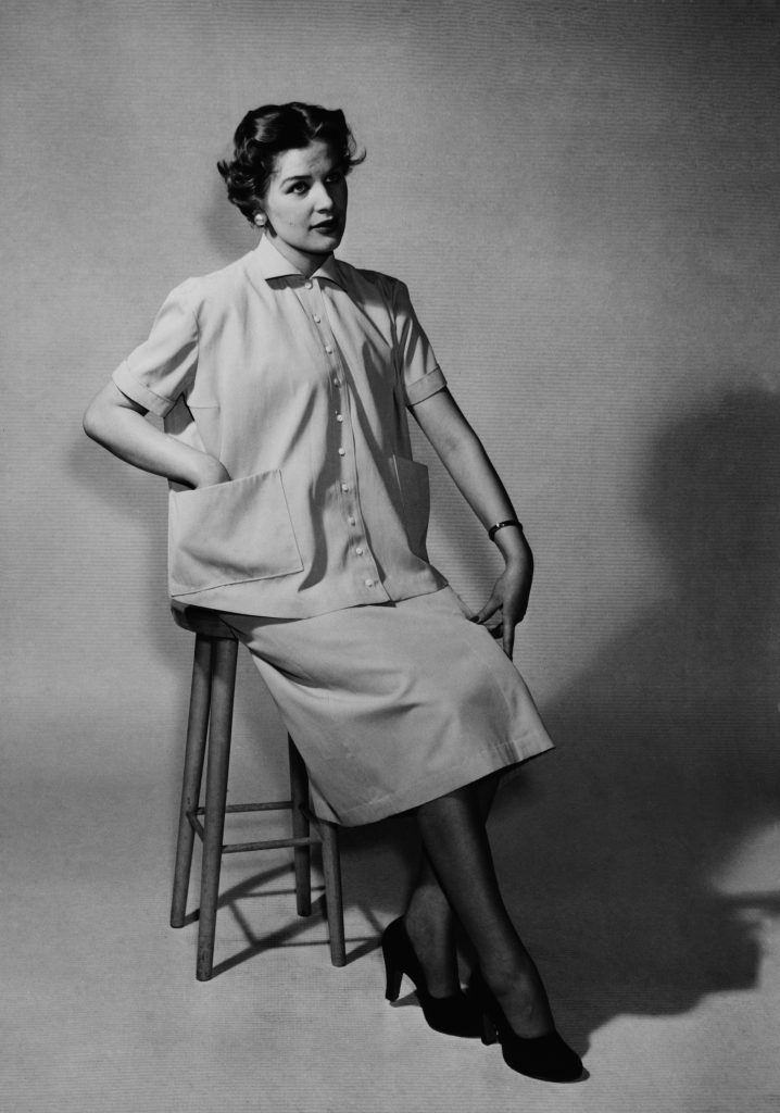 Naismalli esittelee pukua, johon kuuluu lyhythihainen vaalea kauluspaita sekä samansävyinen hame. Nainen istuu studiossa korkealla jakkaralla.