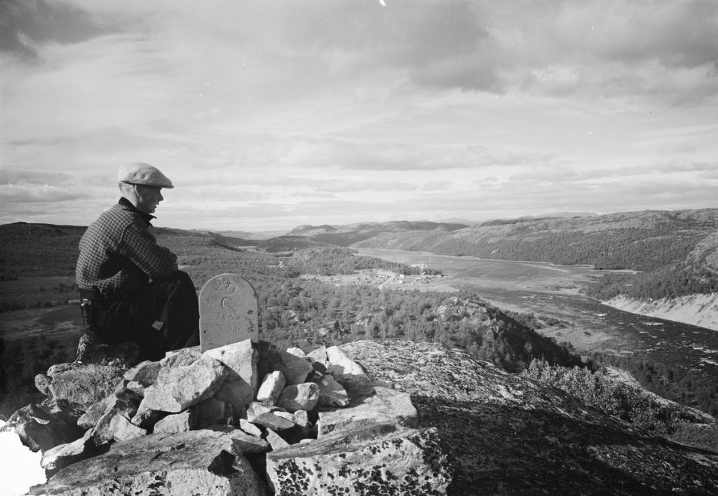Lättähattuun ja flanellipaitaan pukeutunut mies istuu korkean mäen huipulla ja katselee maisemaa, josta erottuvat tunturit ja joki.