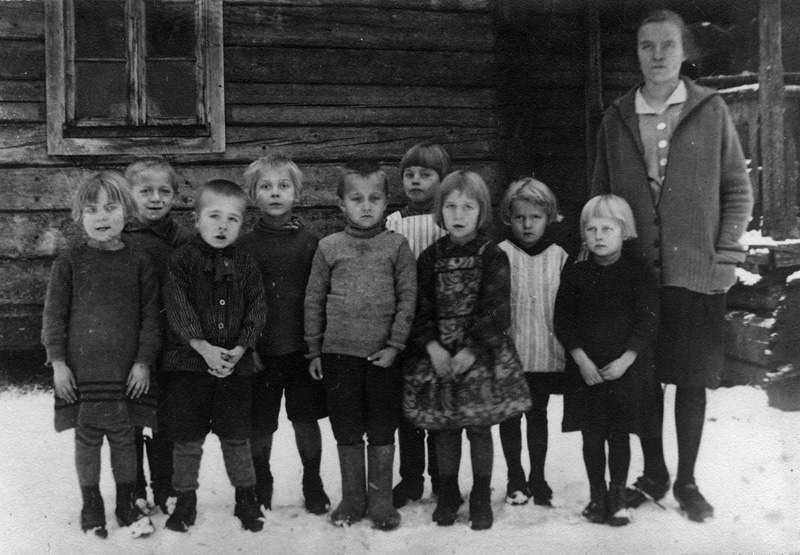 Mustavalkoinen ryhmäkuva yhdeksästä pienestä lapsesta, jotka seisovat ulkona lumessa poseeraamassa kuvaajalle hirsitalon edessä. Kuvassa oikealla seisoo lasten opettaja.