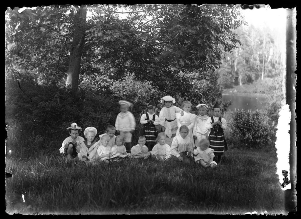 Etualalla nurmikolla istuu yhdeksän lasta, ja heidän takanaan seisoo kuusi lasta. Kaikki lapset katsovat kameraan, ja kaksi heistä piteleen sylissään marsua. Kuvan taka-alalla näkyy pensasaita, suuri lehtipuu sekä vesistöä.
