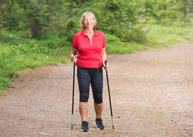 Kuvassa iloinen nainen kävelysauvoilla lenkkeilemässä luonnossa.