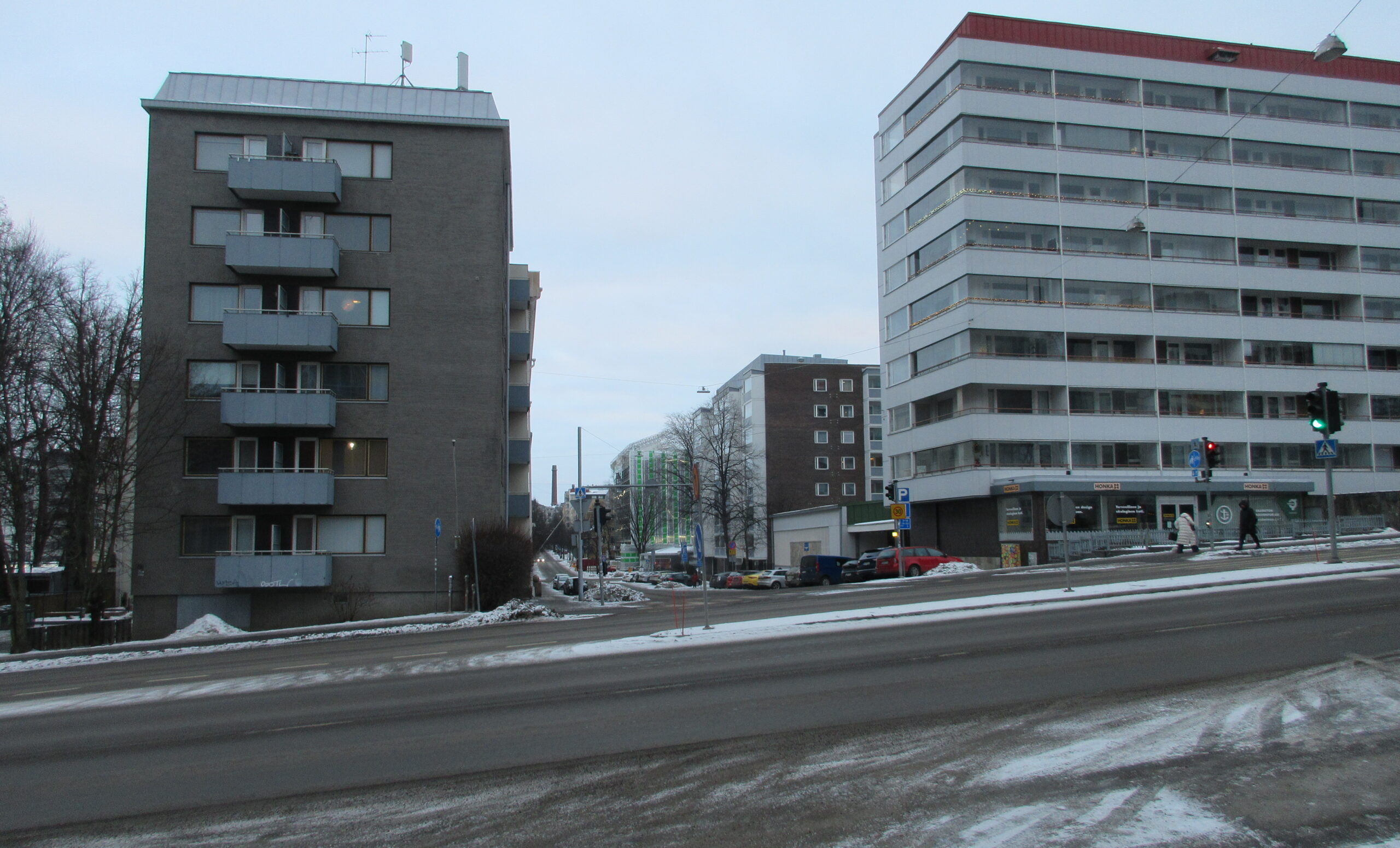 Talvinen, vähäluminen tienäkymä risteyksestä Turussa. Etualalla alamäkeen oikealta vasemmalle kulkeva sula asfalttitie, taustalla kauemmas loittoneva pienempi katu. Loittonevan kadun päässä vasemmalla puolella korkea kerrostalo vasemmalla puolella, samoin kadun oikealla puolella. Loittonevan kadun varrella kerrostaloja, pysäköityjä autoja ja kadun päässä savupiippuja.