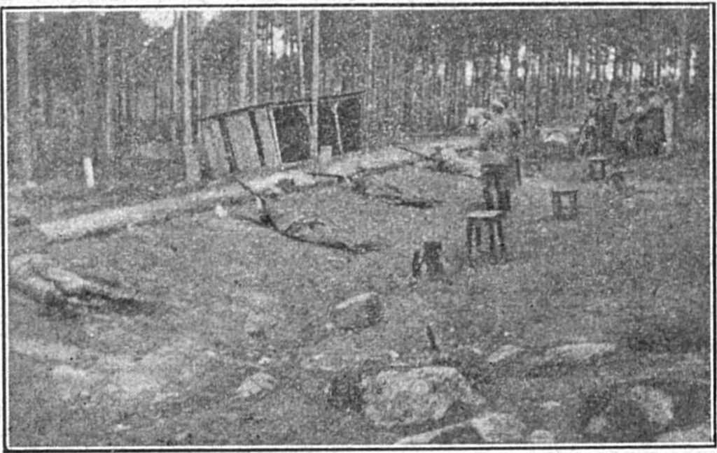 Mustavalkoinen valokuva makaavista miehistä ampumassa kivääreillä maavallin takana. Ampujien takana kaksi seisaallaan seuraavaa miestä, taustalla näkyy laudoista rakennettu puuvaja.
