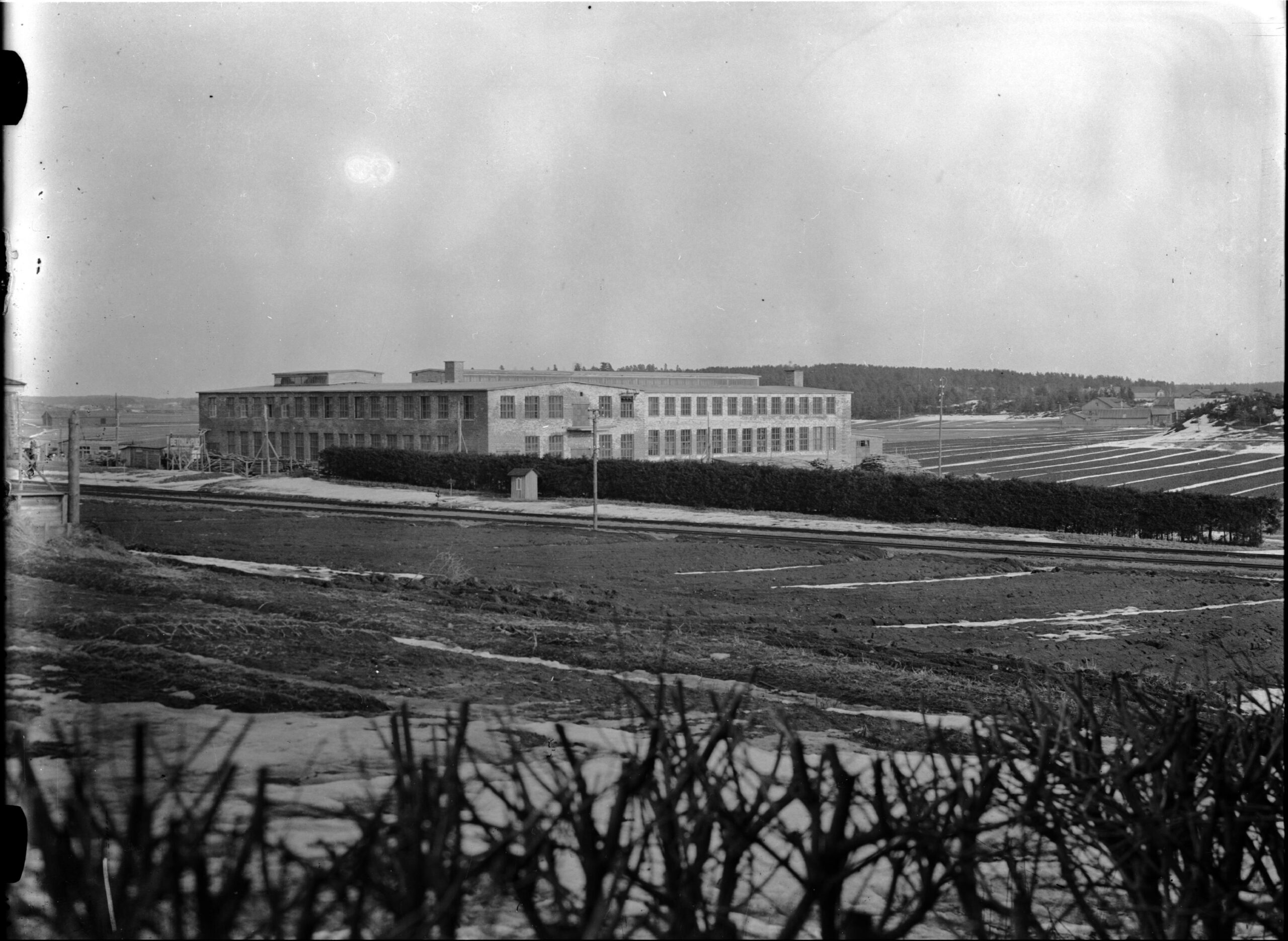 Suuri tiilinen tehdasrakennus, Tolfanin tehtaat, peltojen välissä. Tehtaan ikkunat ovat kahdessa rivissä, sen etualalla kulkee maantie sekä puhelinlinja tolppien varassa.