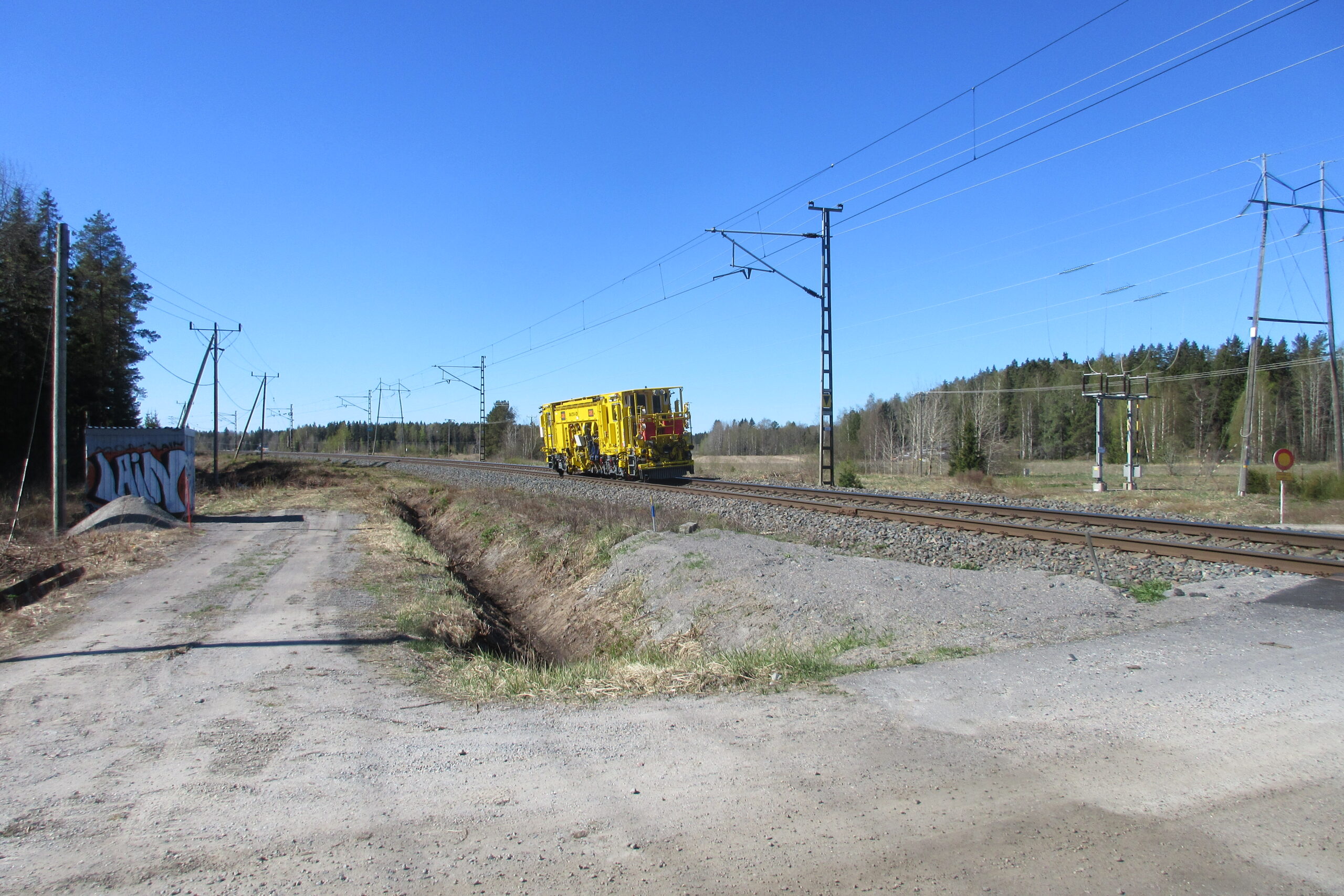 Junaratojen korjaamiseen käytetty keltainen työkone, raiteentukemiskone ajamassa raiteilla lähellä ylikulkukäytävää.